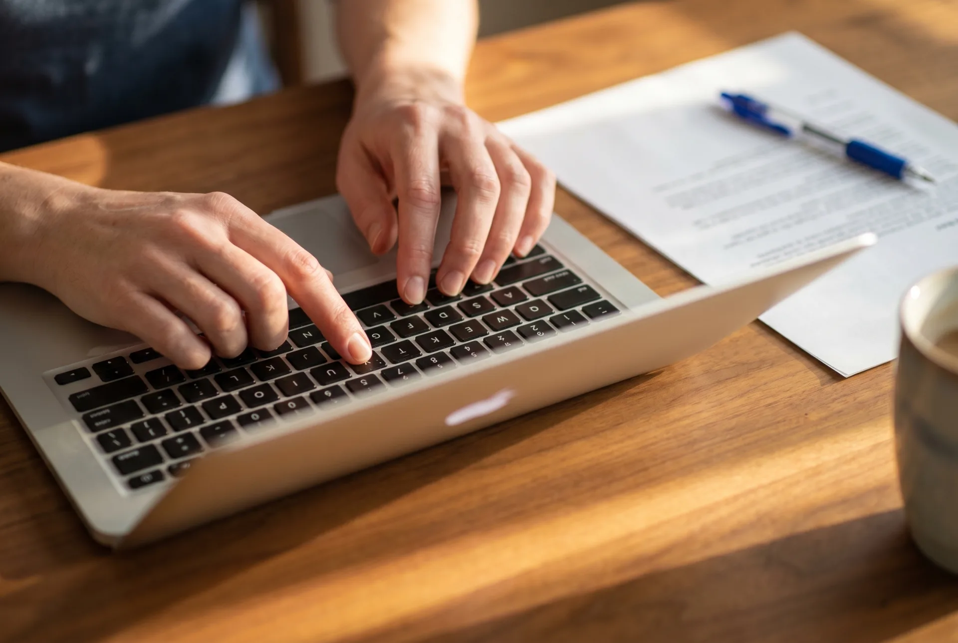 Close-up of hands typing on a silver laptop keyboard with a printed letter template and a blue ballpoint pen visible on the warm wooden desk beside the laptop in soft morning window light