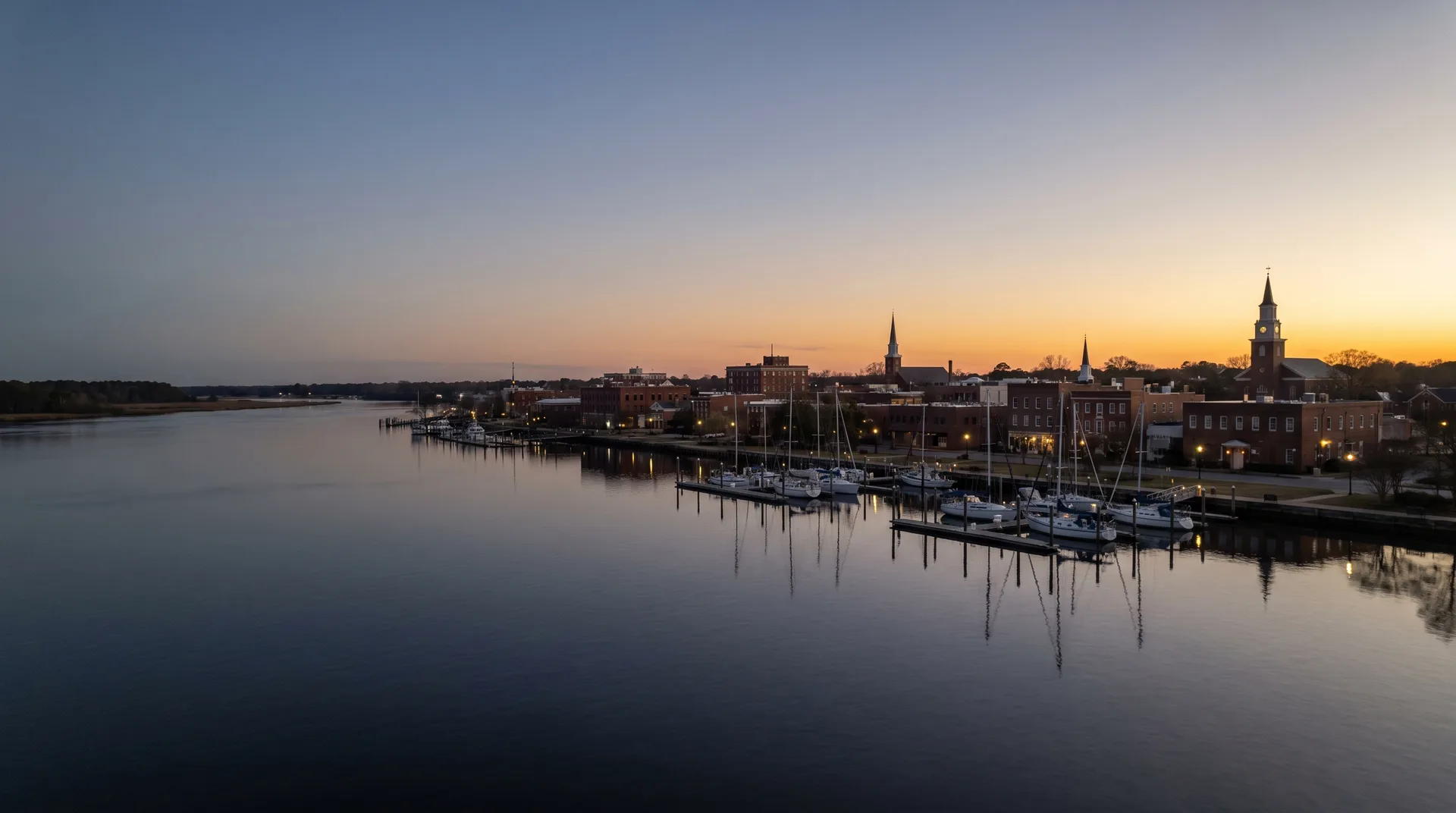 The New Bern waterfront at dusk, church steeples and brick buildings beside a marina on the river